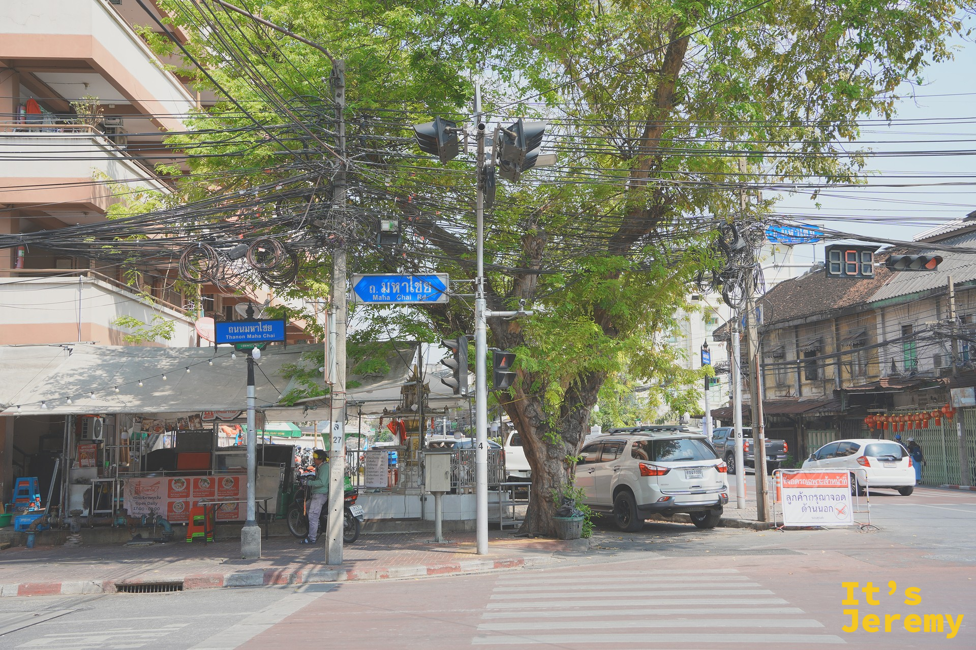 另一個角度的 Mahachai 路口街景，藍底路牌與綠樹下的老城區午後