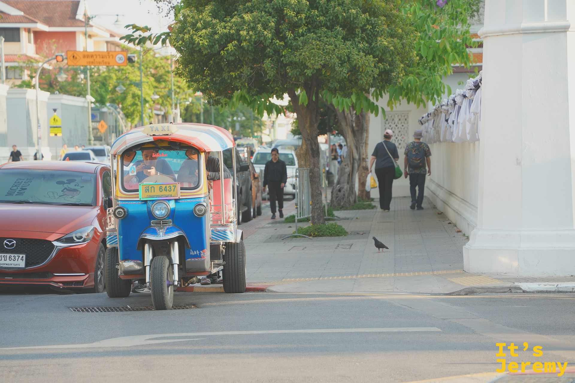 一輛藍色 tuk-tuk「ส 6437 TAXI」從紅色 Mazda 旁邊開過、遠處走著一對老夫妻——通往 Tha Tien 碼頭最後 200 公尺