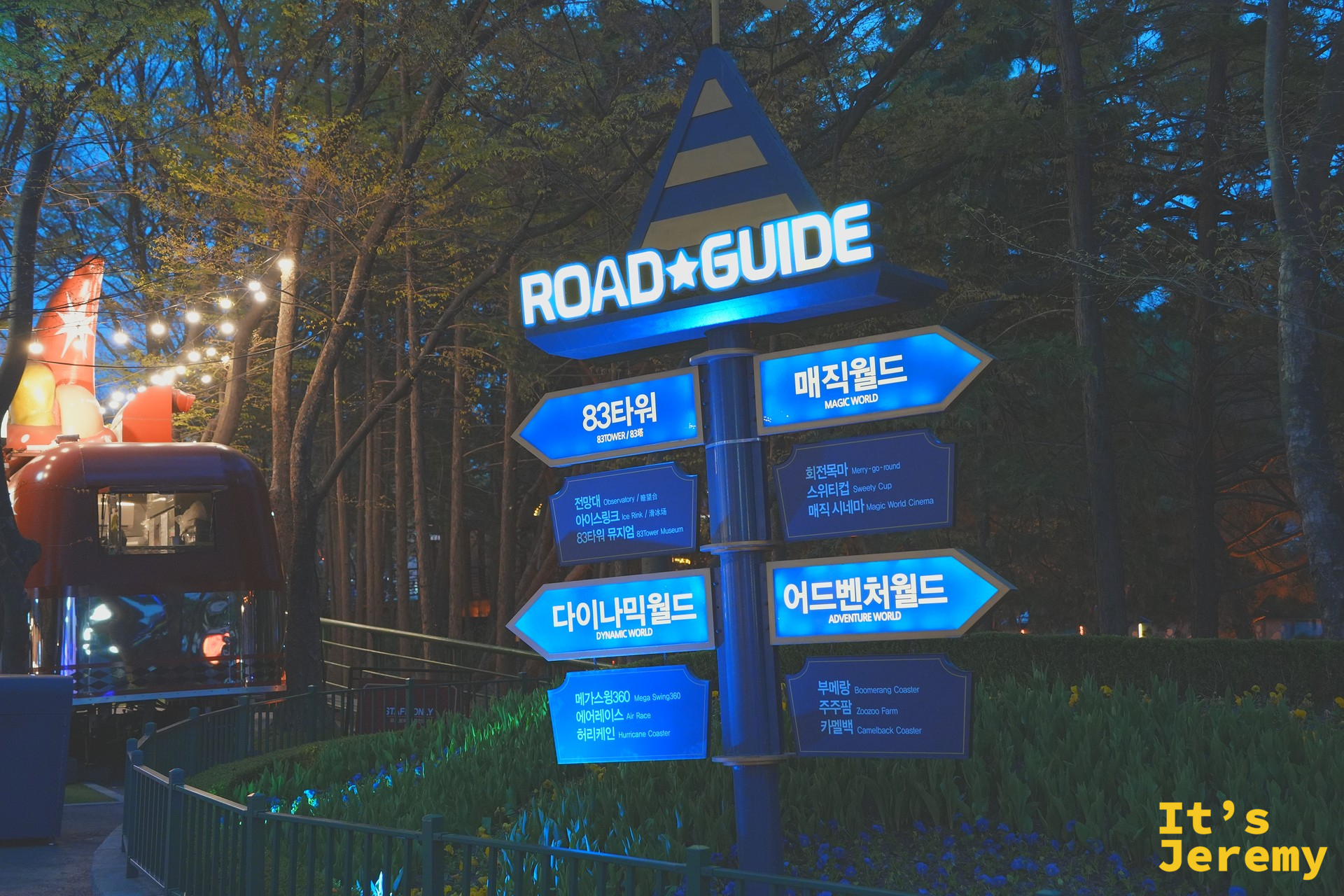 園區內的 Road Guide 藍色四向路標,最上方是三角形「ROAD ★ GUIDE」標示,下方四面藍色路標分別指向「83타워」、「매직월드」、「다이나믹월드」、「어드벤처월드」四個分區,路標座落在一片紫色花圃上