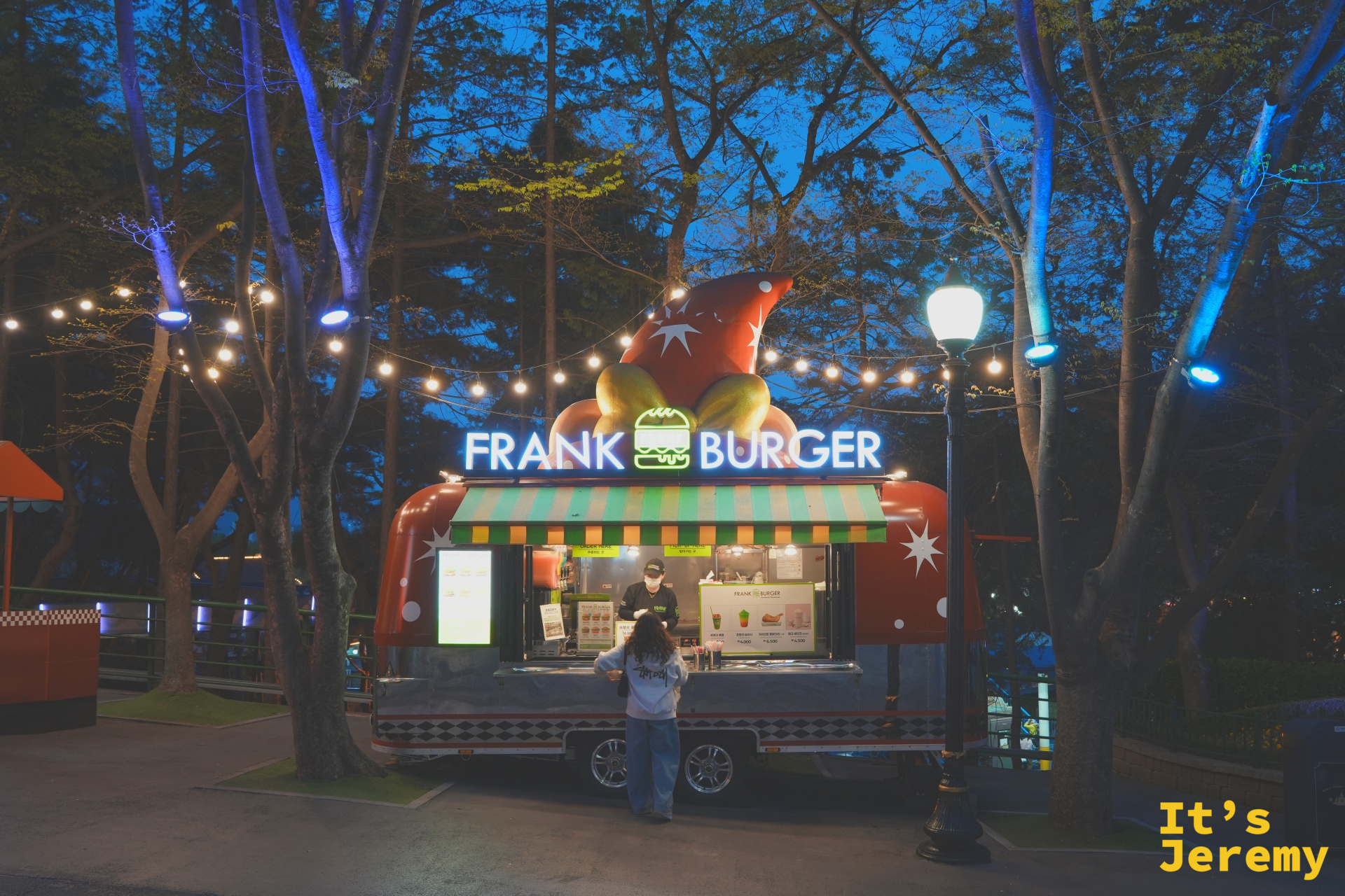 Frank Burger 餐車,紅色木屋造型車身、車頂是漢堡蔬菜造型雕塑,白底霓虹「FRANK BURGER」招牌,前方掛著一串黃色小燈泡,一位戴帽子的人站在窗口前