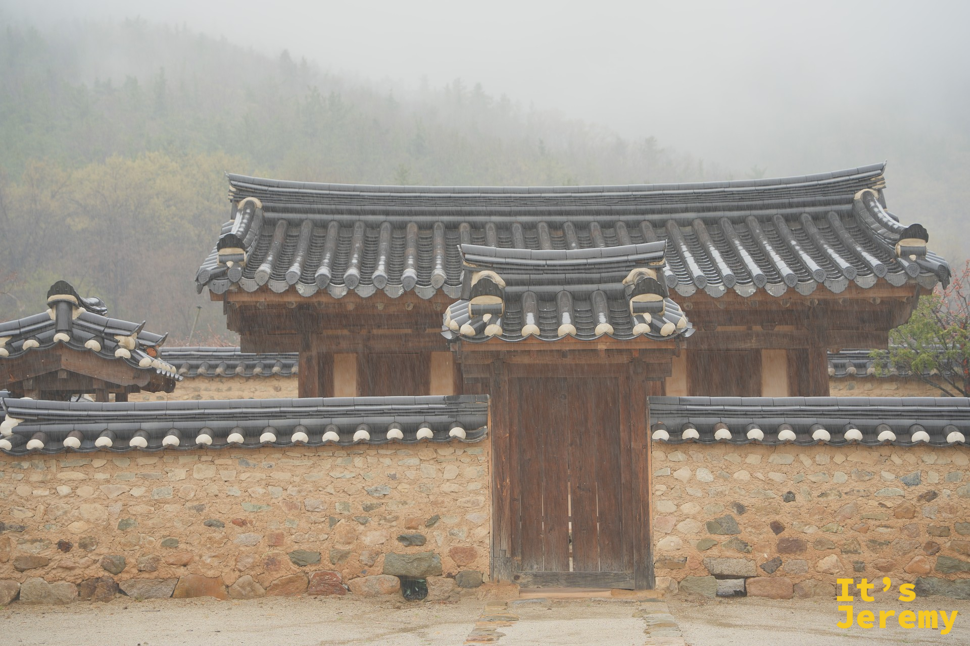 雨中的漆谷韓屋村正門,青瓦大門後是霧氣瀰漫的山,雨絲清晰可見