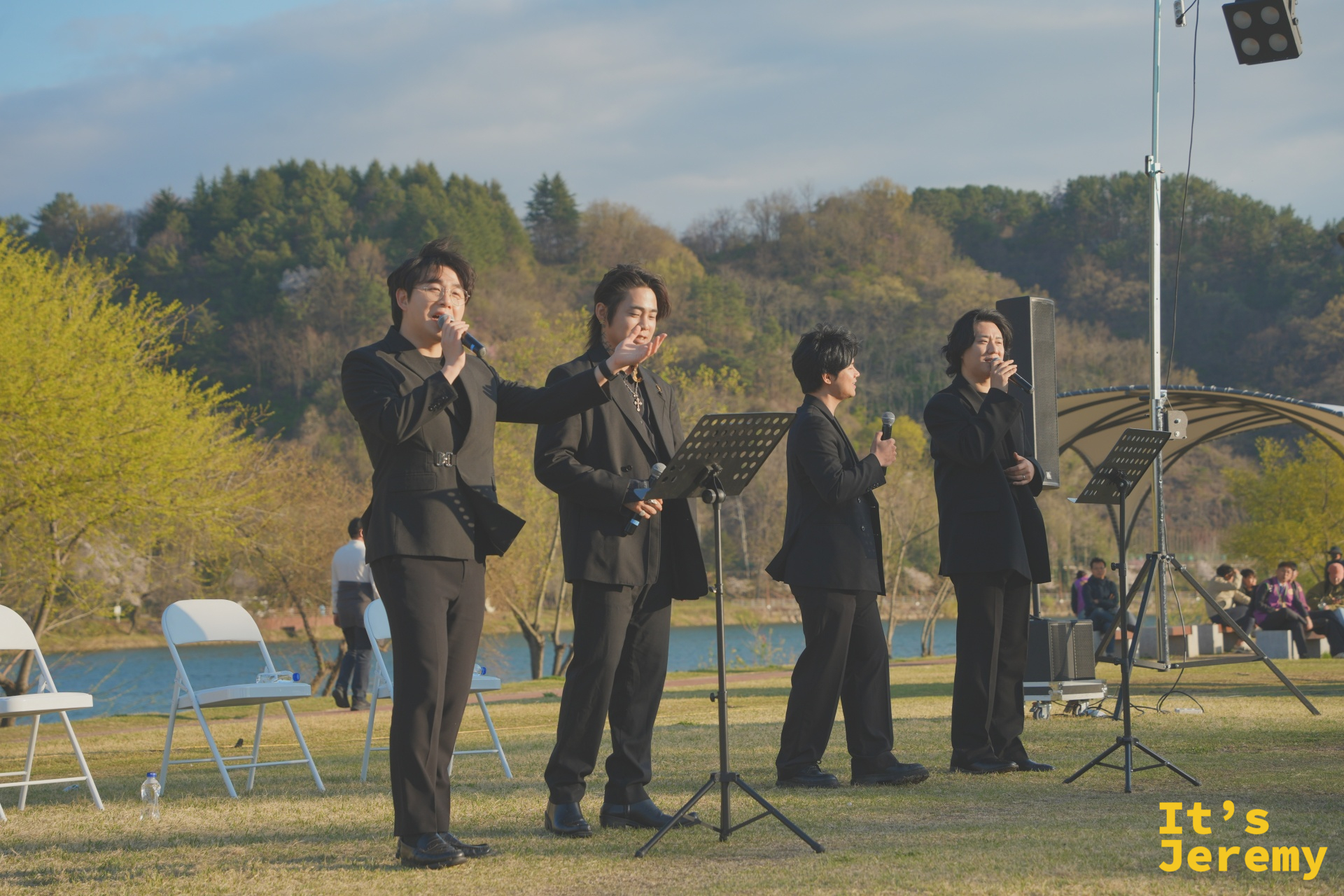 四名男聲組合穿黑西裝在江邊草地演出,背景是寬闊的新安川江面與對岸山丘