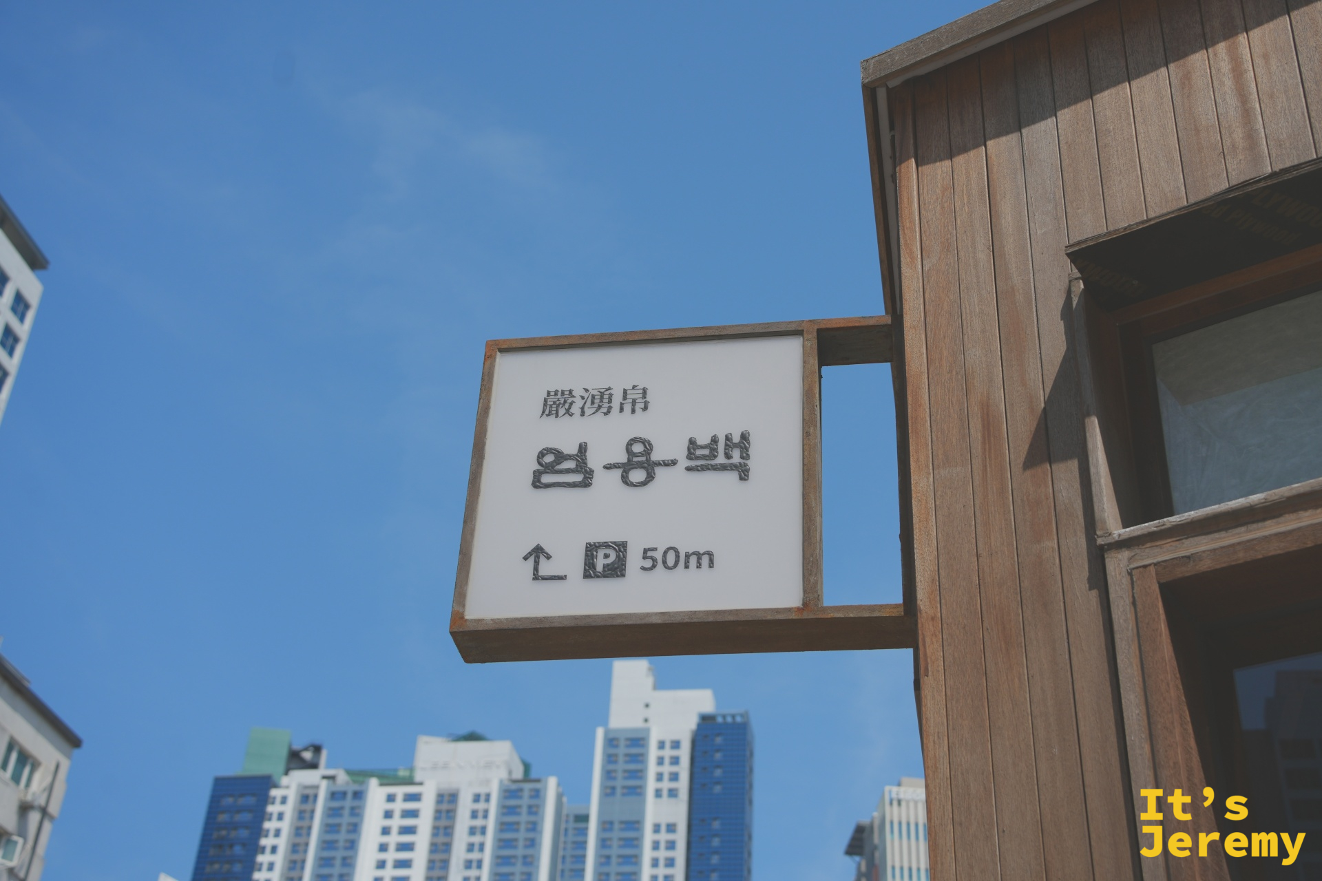 店外路標指示,漢字「嚴湧帛」清晰可見,藍天下簡潔俐落的木框招牌